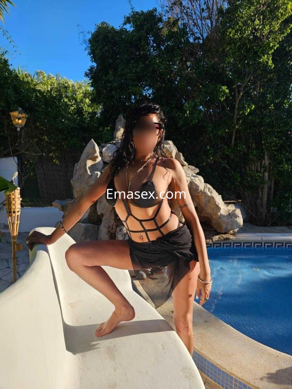 La puta posa en un entorno exterior junto a una piscina, luciendo un bikini negro y una falda transparente.