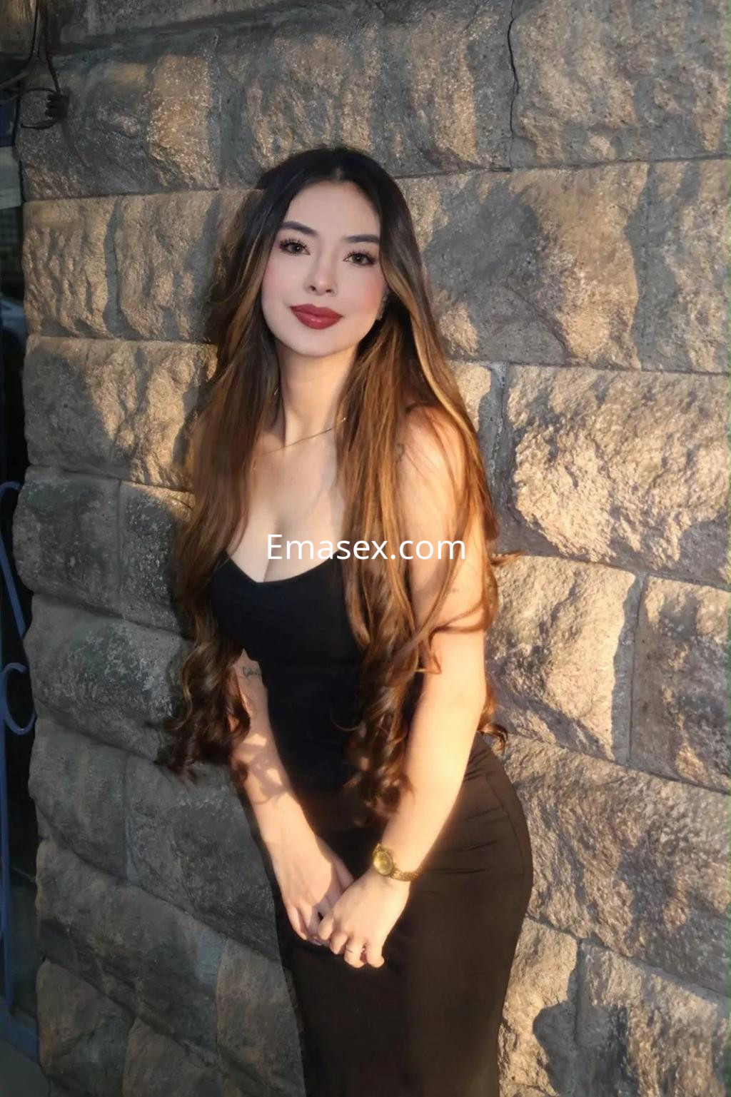 Puta de cabello largo y ondulado, vestida con un ajustado vestido negro, posando junto a una pared de piedra en Oviedo.