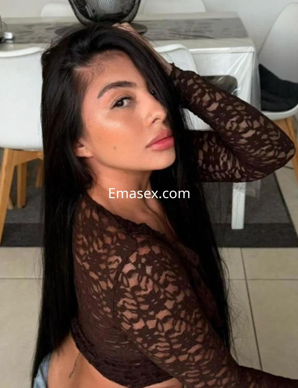 Escort en Madrid con cabello largo y lacio, vistiendo una blusa de encaje marrón, posando de lado en un ambiente luminoso.