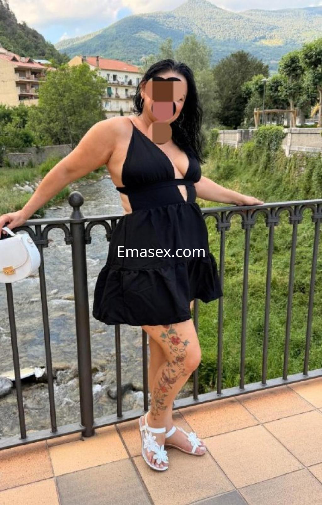 La puta luce un vestido negro con detalle en la cintura y sandalias blancas, posando junto a un río en Cáceres.