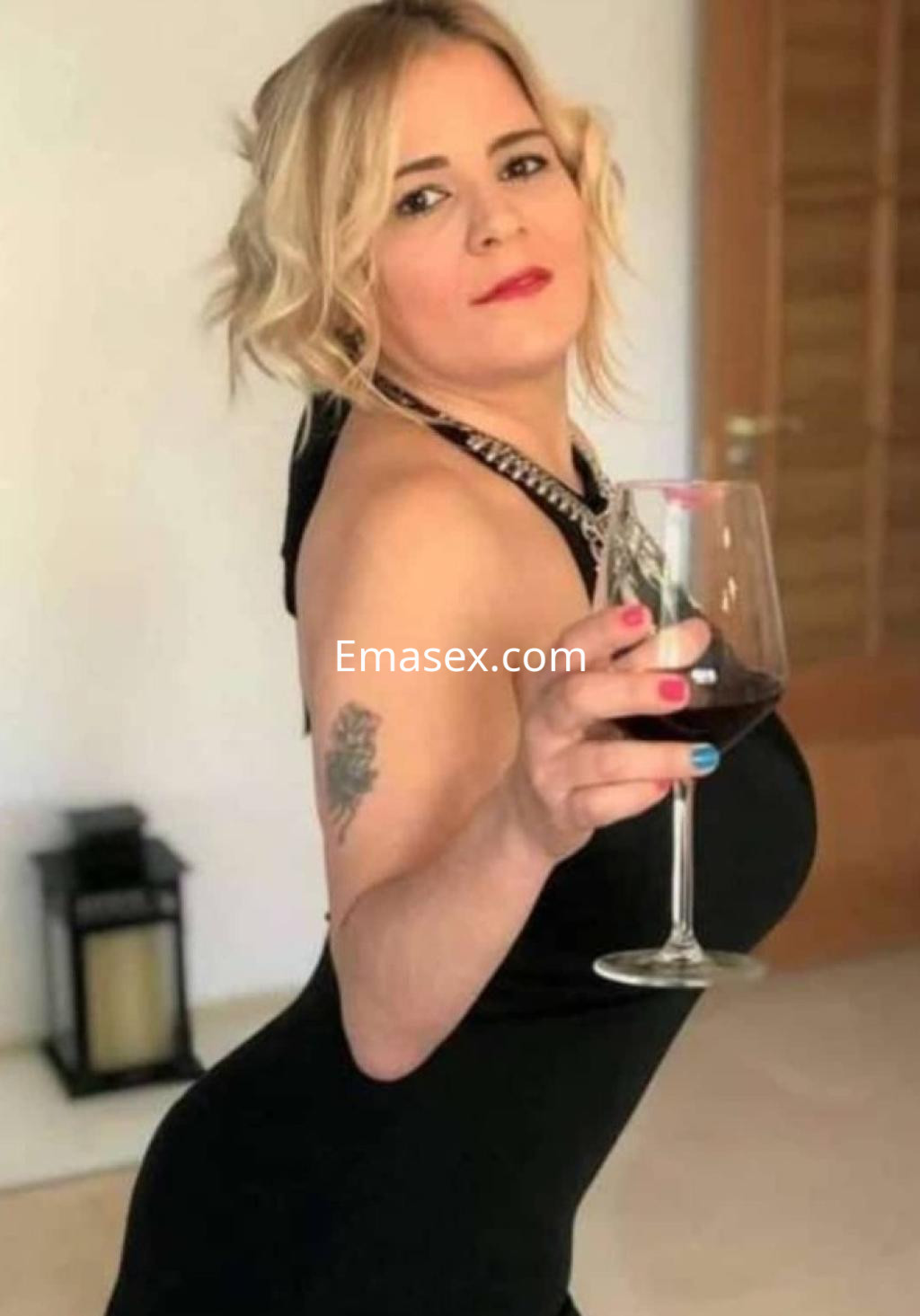 Puta en Pamplona, posando con una copa de vino, lleva un vestido negro ajustado y tiene cabello rubio ondulado.