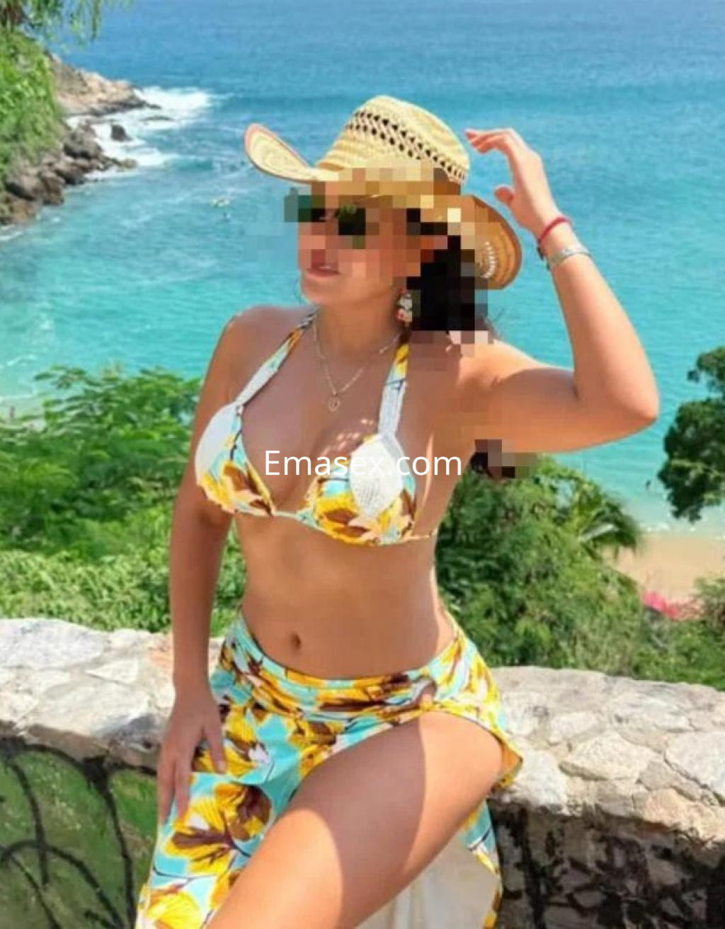 Escort en Albacete vistiendo un bikini colorido y un sombrero, posando junto a un mar azul y paisaje natural.