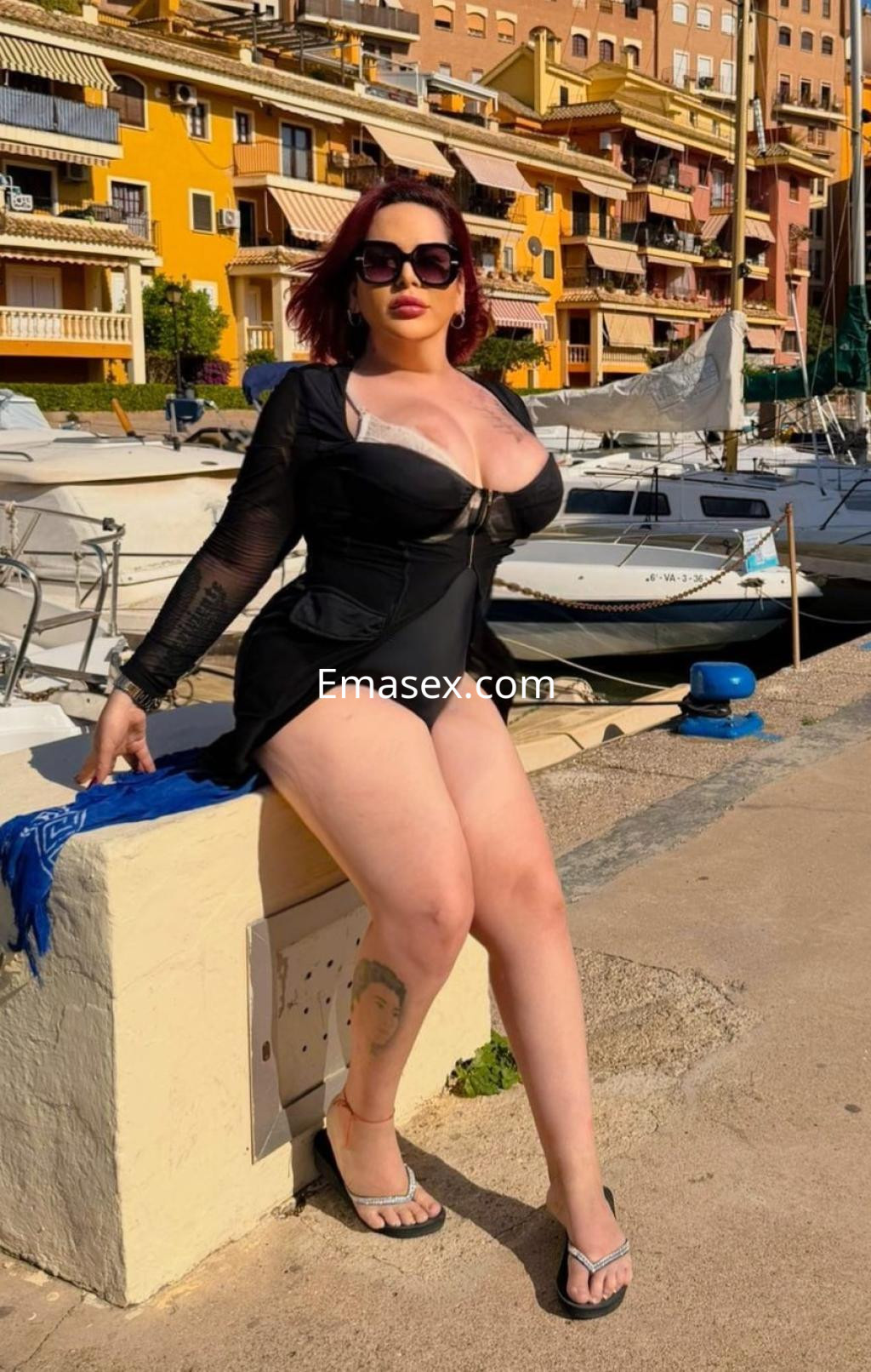 Escort en Alicante vistiendo un traje de baño negro con una chaqueta transparente, posando junto a embarcaciones en el puerto.
