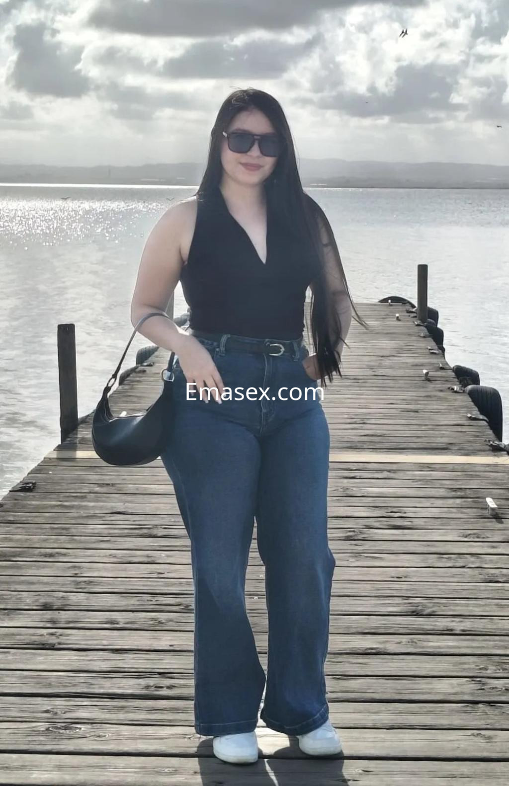 Escort en Alicante posando en un muelle, viste una blusa sin mangas negra y jeans acampanados, con gafas de sol.