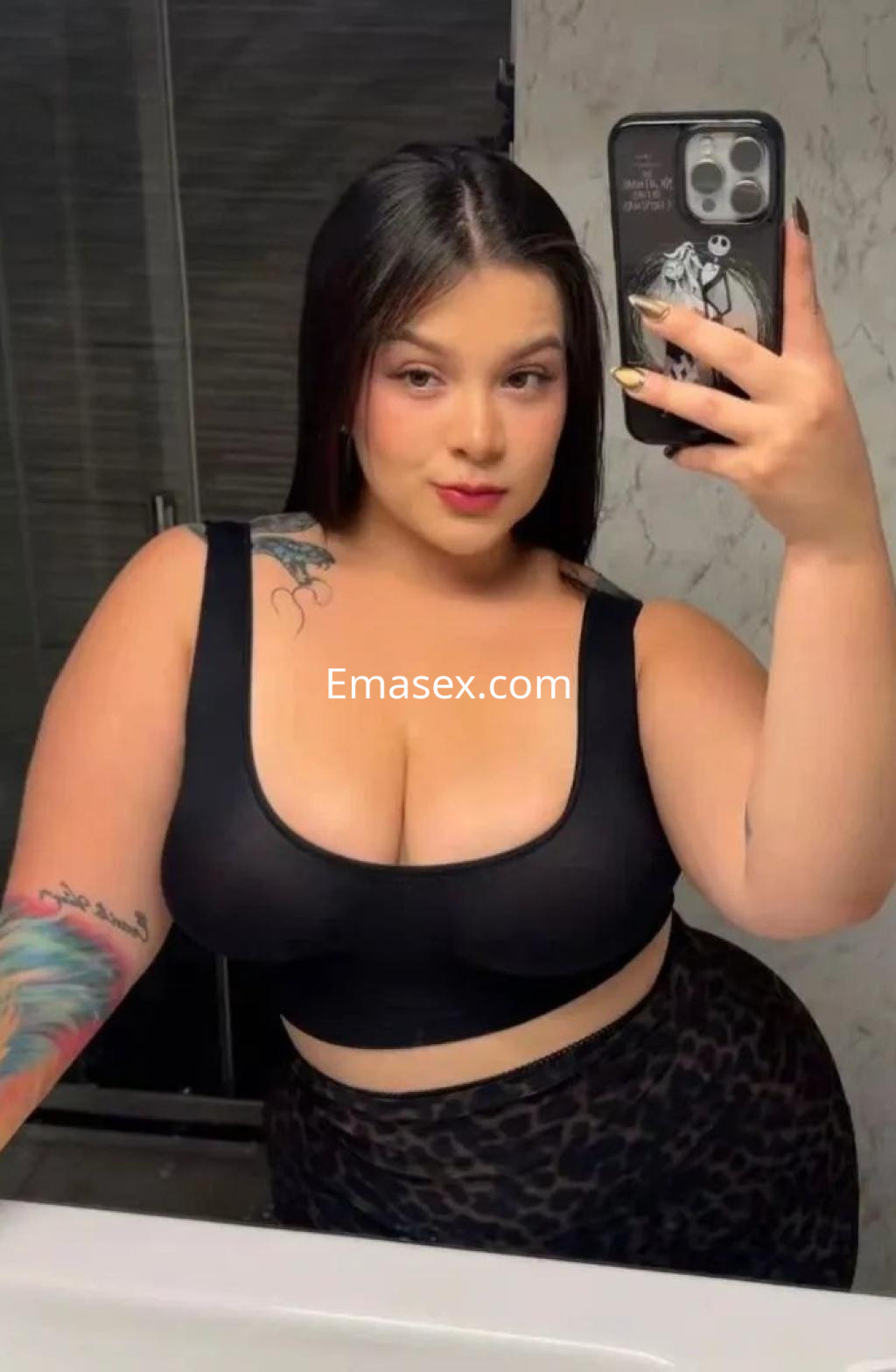 Escort en Alicante en un baño, luciendo un top negro ajustado y una falda de leopardo mientras se toma un selfie.