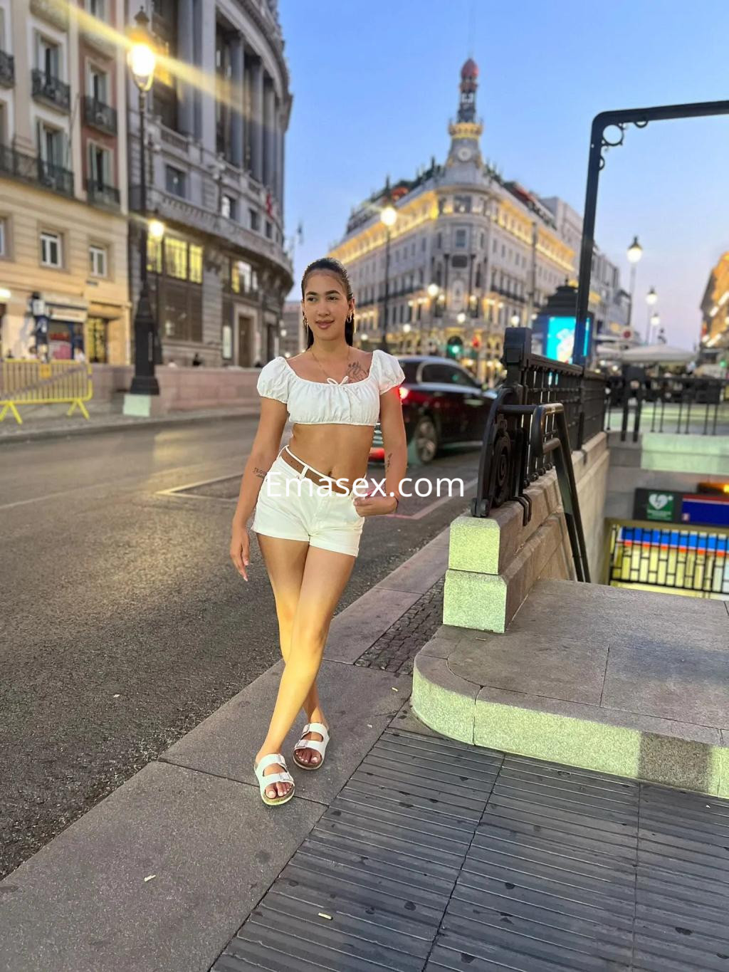 Escort en Vigo, luciendo un top blanco y shorts cortos, posa frente a un entorno urbano iluminado al atardecer.