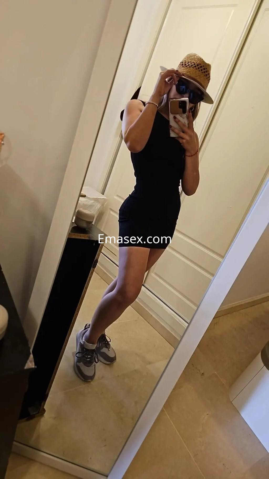 Puta en Málaga posando frente a un espejo, con un vestido negro ajustado, sombrero y zapatillas deportivas.
