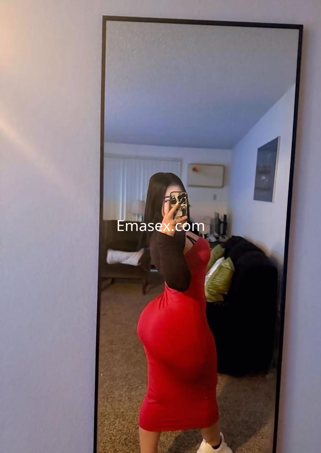 Escort en Irún posando de lado frente a un espejo, vistiendo un vestido rojo ajustado y una blusa negra.