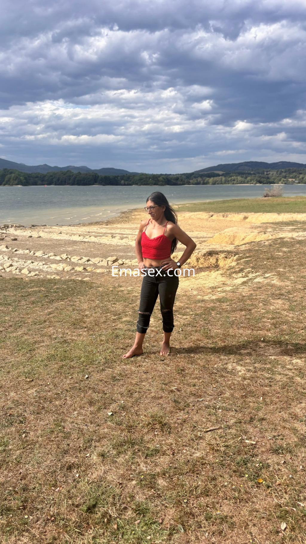 Escort en Irún con blusa roja y pantalones negros, posando descalza en un entorno natural junto al agua.