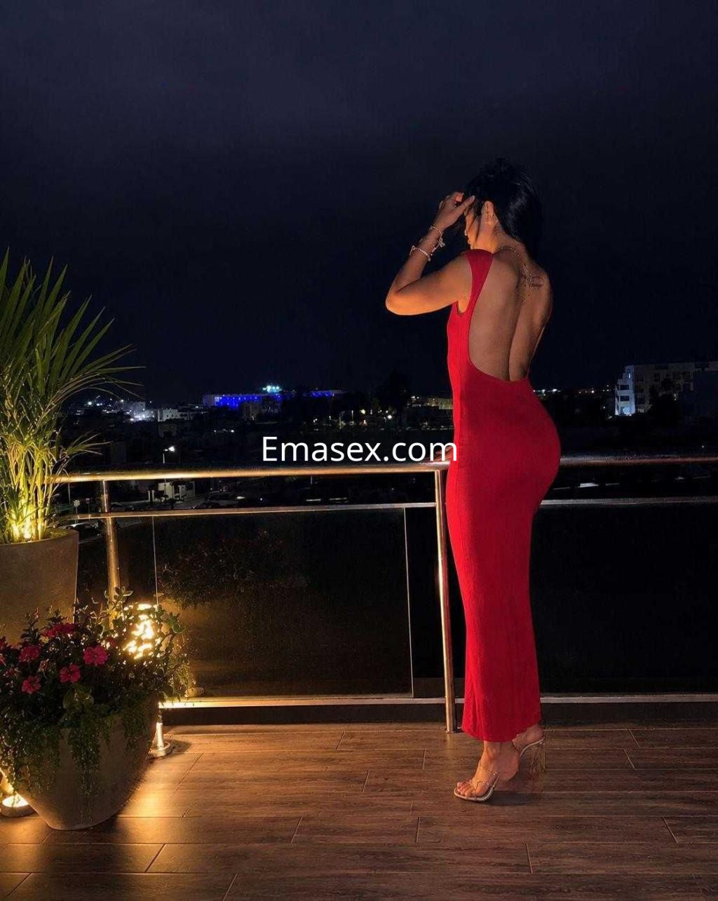 Puta con vestido rojo ajustado y escote en la espalda, posando en un balcón de Ibiza por la noche.
