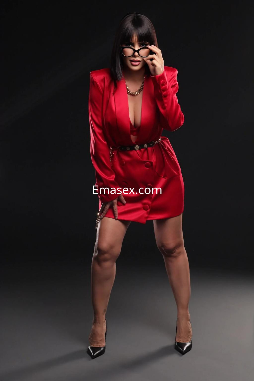 Escort en Oviedo viste un vestido rojo con escote, tacones negros y gafas, posando de forma seductora.