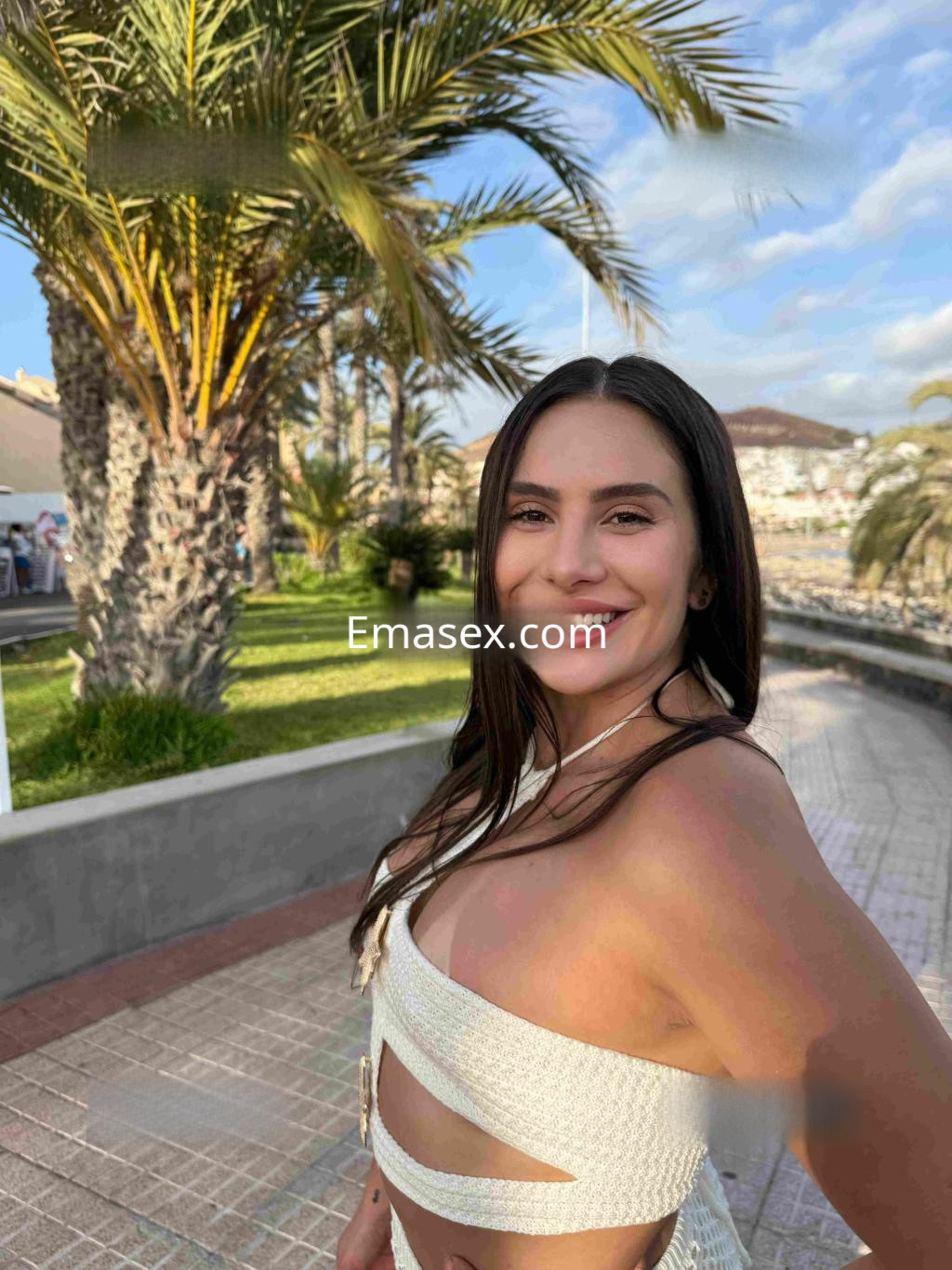 Escort sonriente en Tenerife, luciendo un vestido blanco sin mangas, con palmeras al fondo y una atmósfera vibrante.