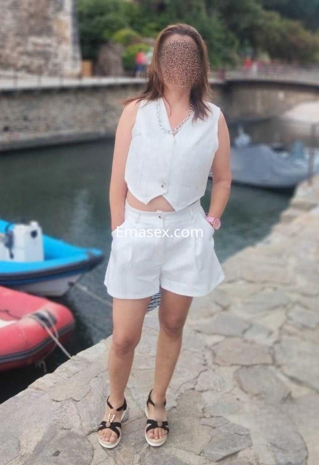 Puta en Zaragoza de pie junto al agua, vistiendo un conjunto blanco de top y pantalones cortos, con sandalias.