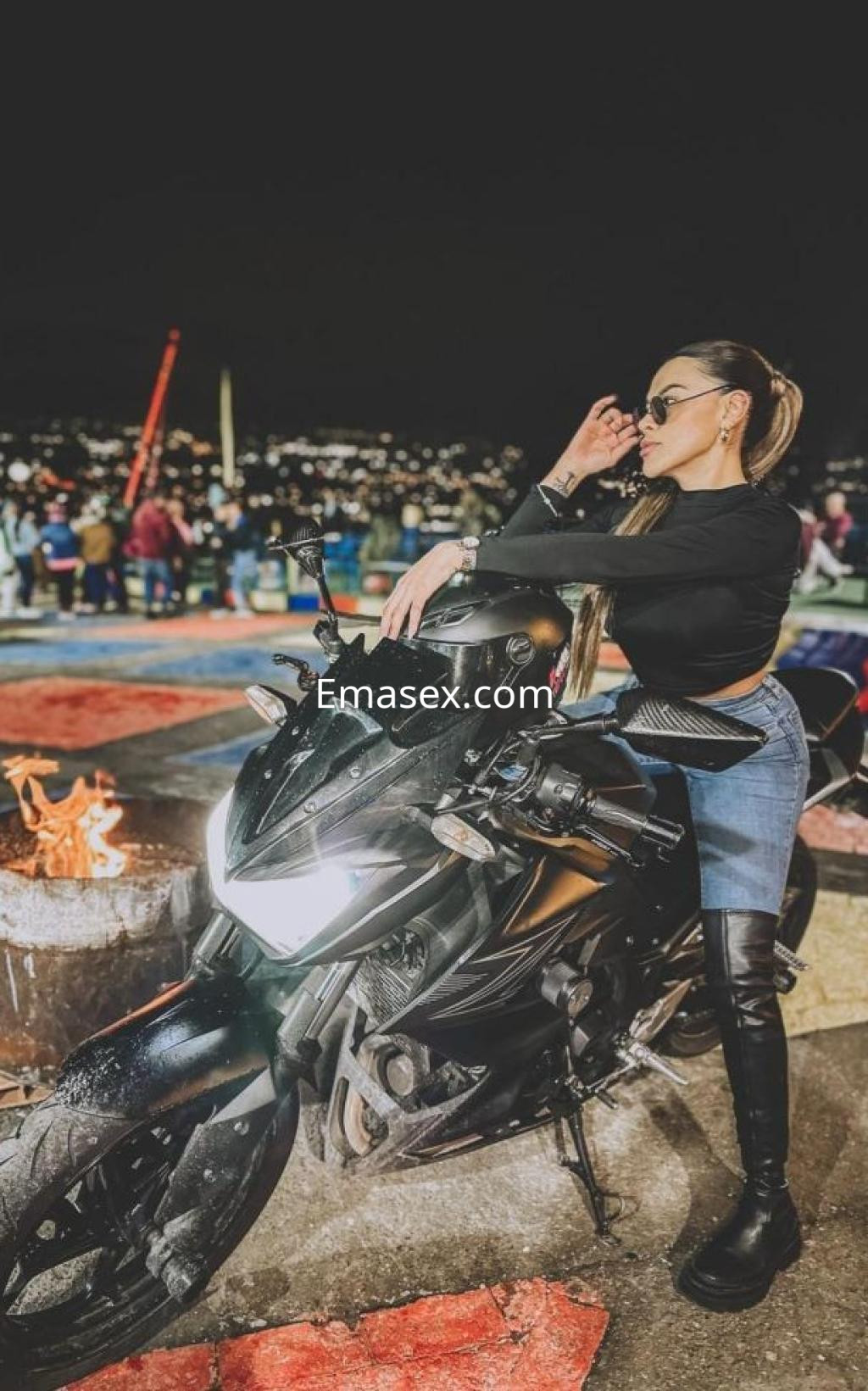 Puta posando sobre una moto negra en Castellón, con un top negro, jeans ajustados y botas largas, luciendo gafas de sol.