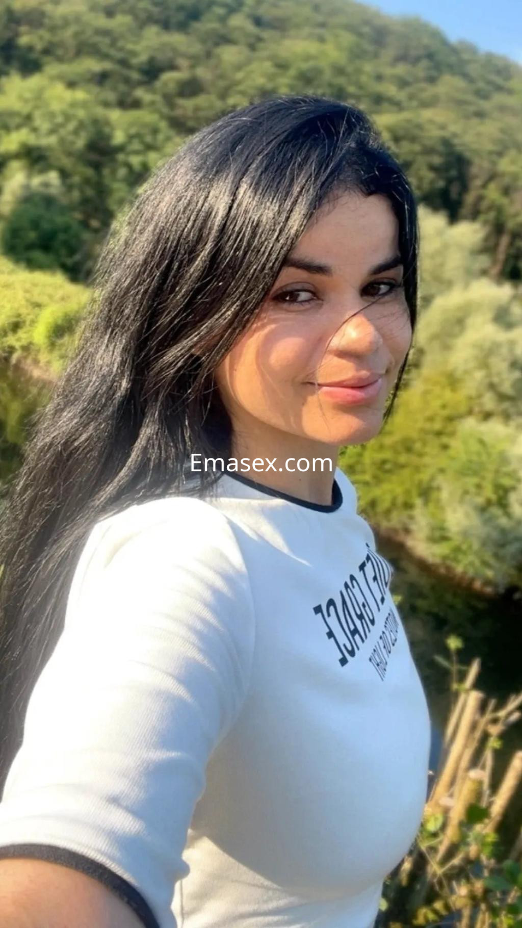 Puta en Coruña, con cabello largo y oscuro, viste una camiseta blanca con detalles en negro, sonriendo en un entorno natural.
