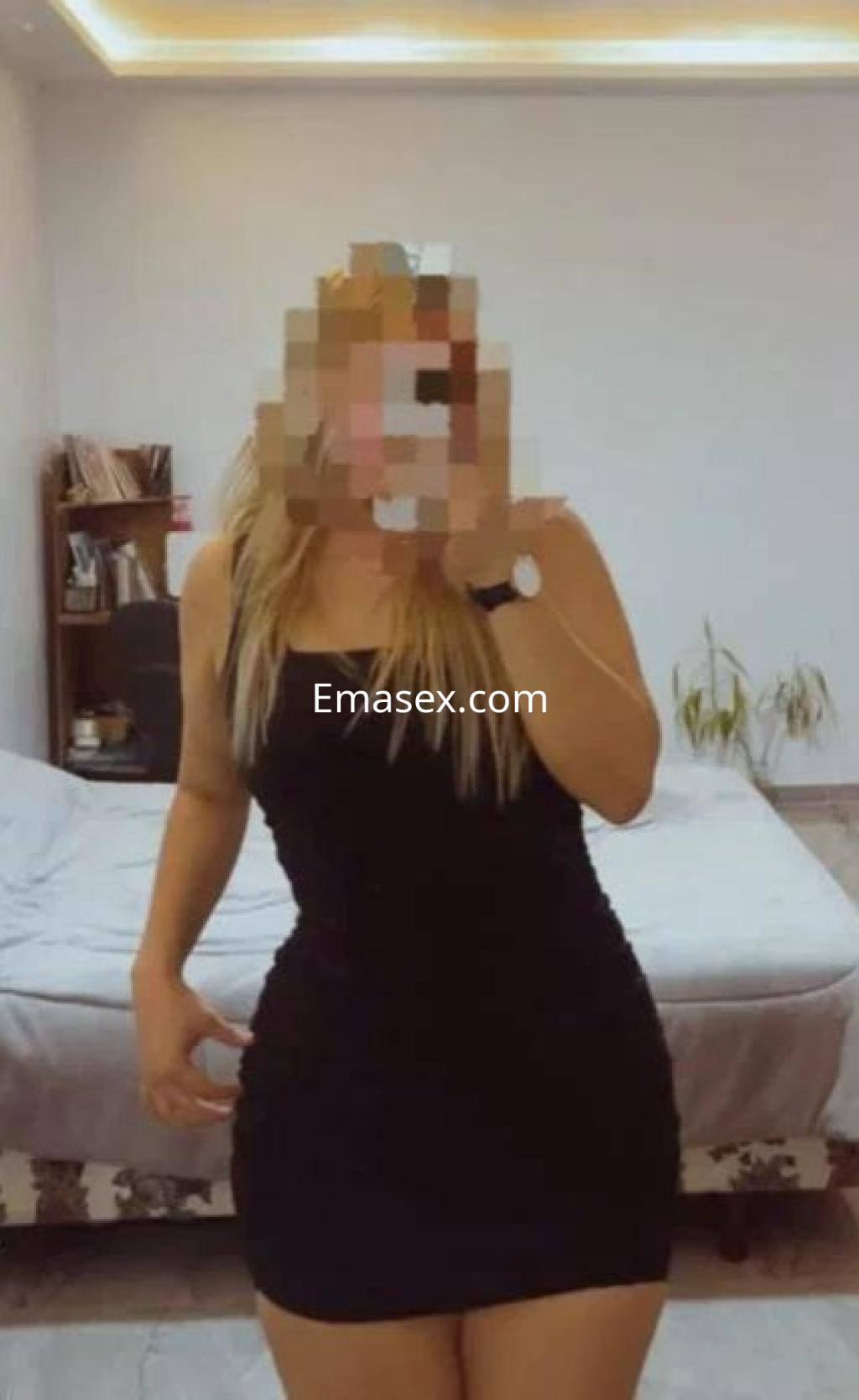 Puta en Cáceres luciendo un vestido negro ajustado, posando frente a una cama en un ambiente luminoso.