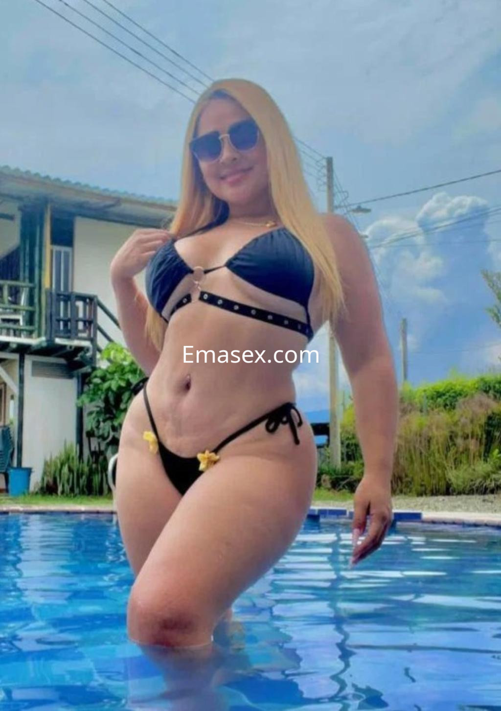 Puta en Valencia, luciendo un bikini negro con detalles en amarillo mientras posa en una piscina al aire libre.