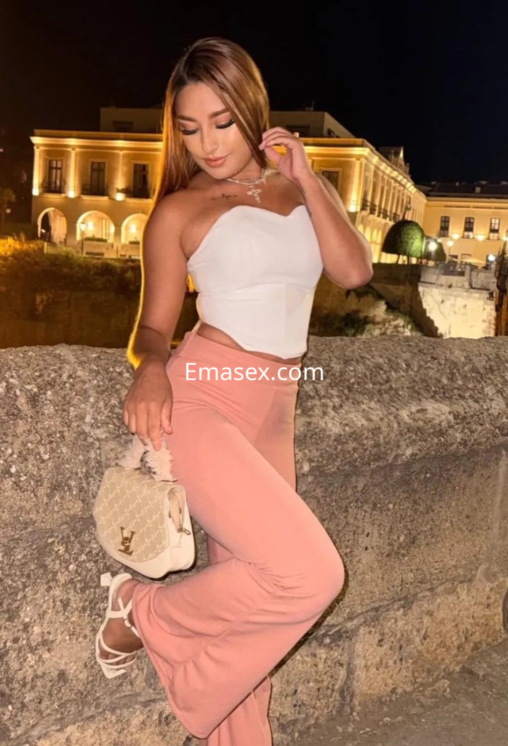 Escort en Alicante con cabello largo y lacio, usando un top blanco y pantalones rosa, posando en un entorno nocturno.