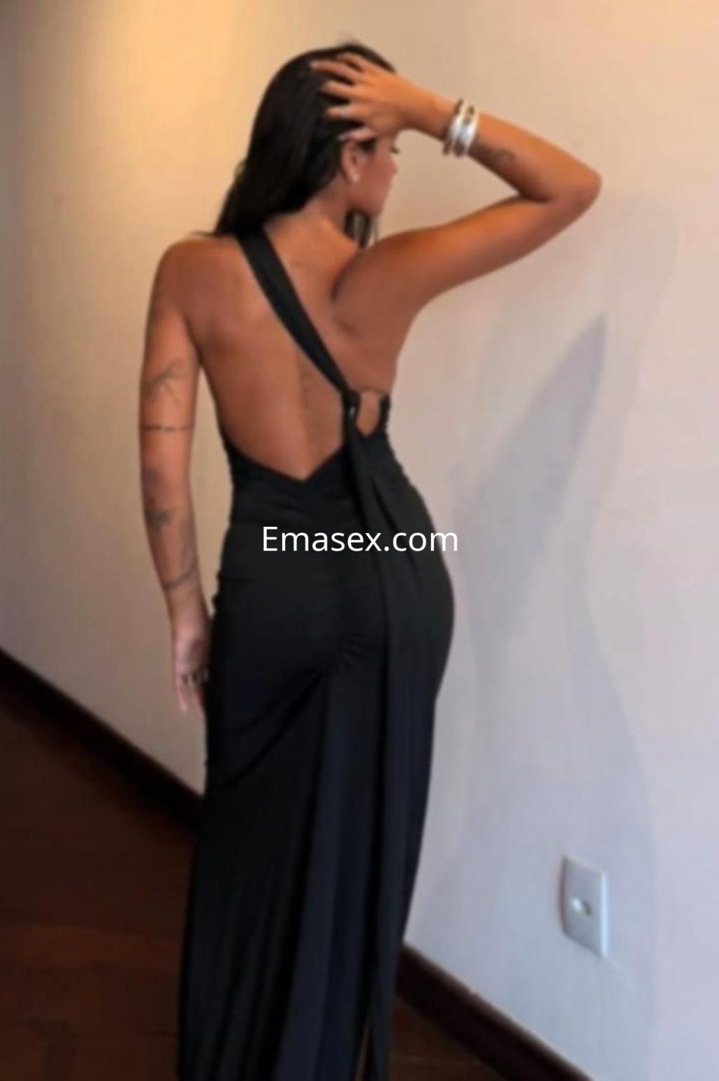 Puta en Alicante, vistiendo un vestido largo negro de corte asimétrico, posando de espaldas en un interior elegante.