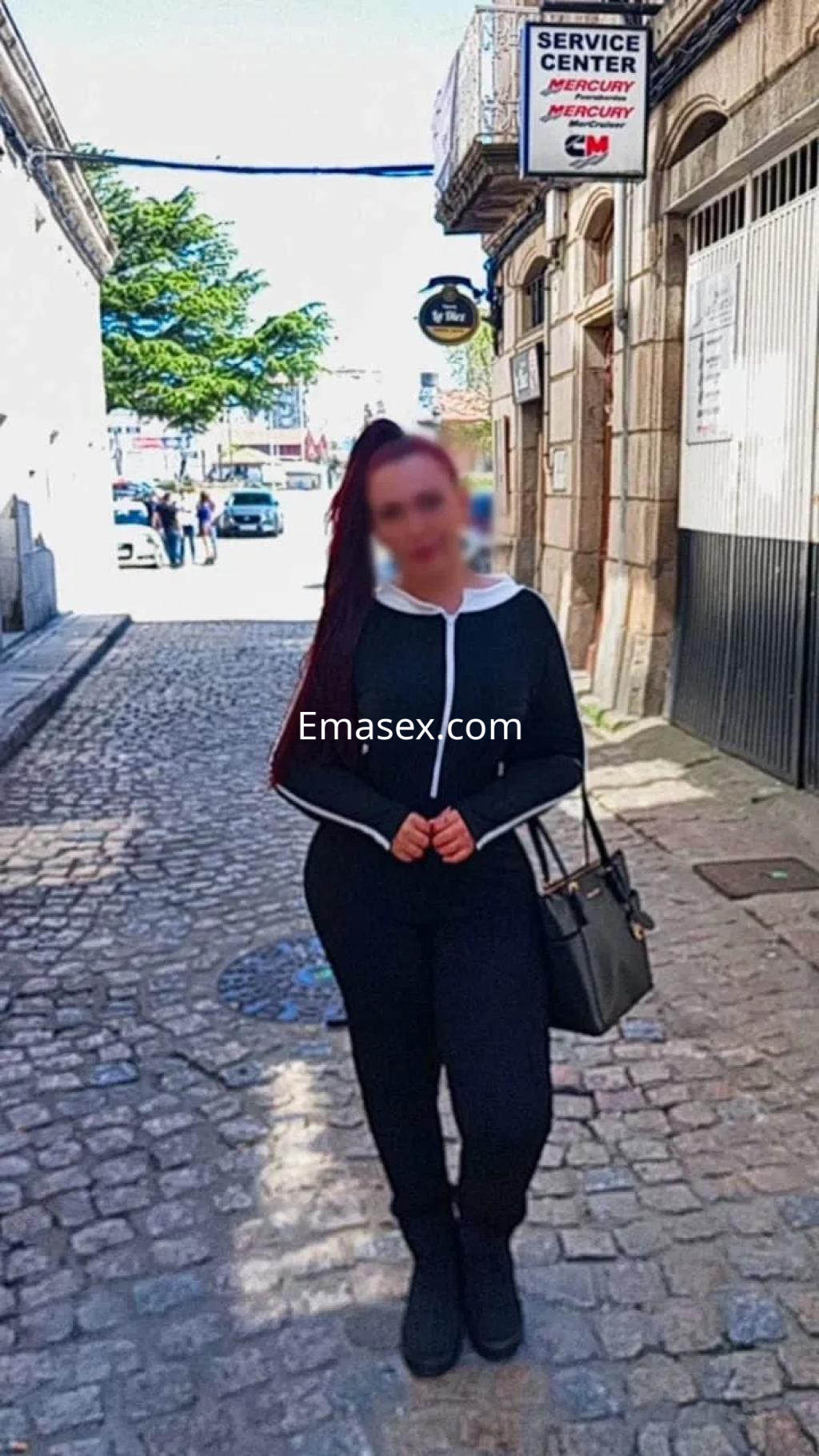 La escort en Vigo se presenta en una calle empedrada, vistiendo un mono negro y llevando un bolso.