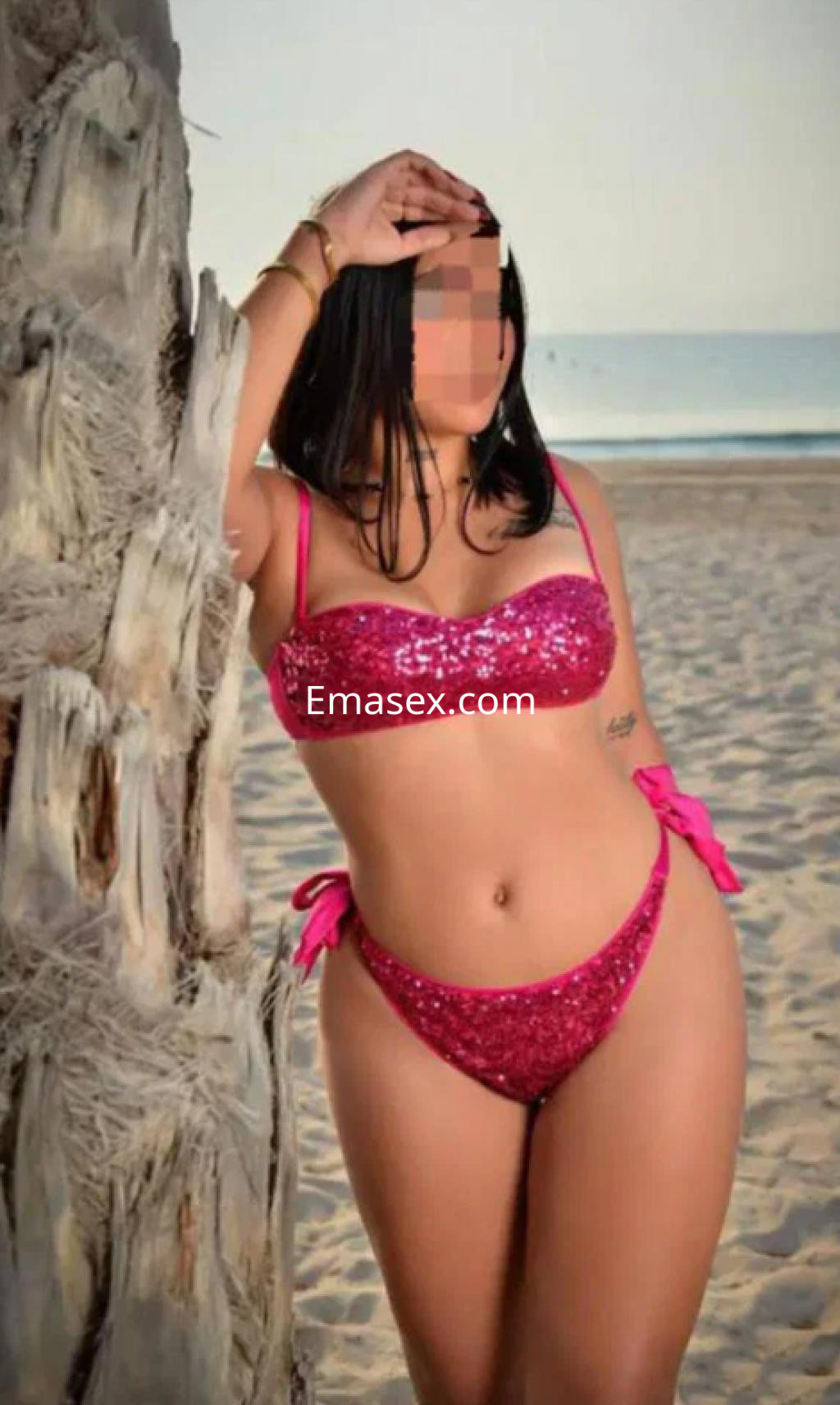 Puta en Albacete con bikini rosa brillante frente al mar, posando junto a una palmera en la playa.