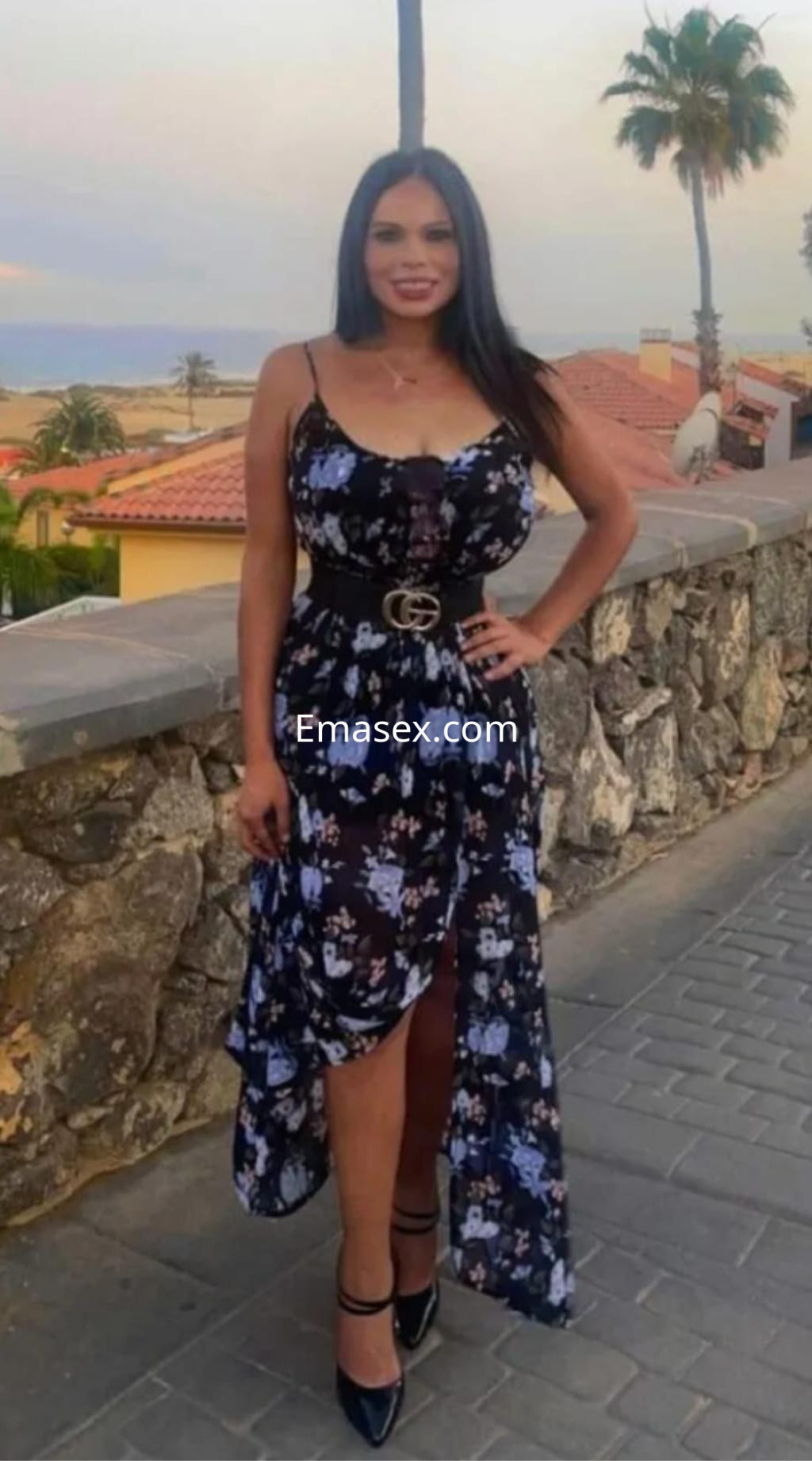 Puta trans en Irún con un vestido largo de flores, posando con una mano en la cintura frente a un paisaje costero.