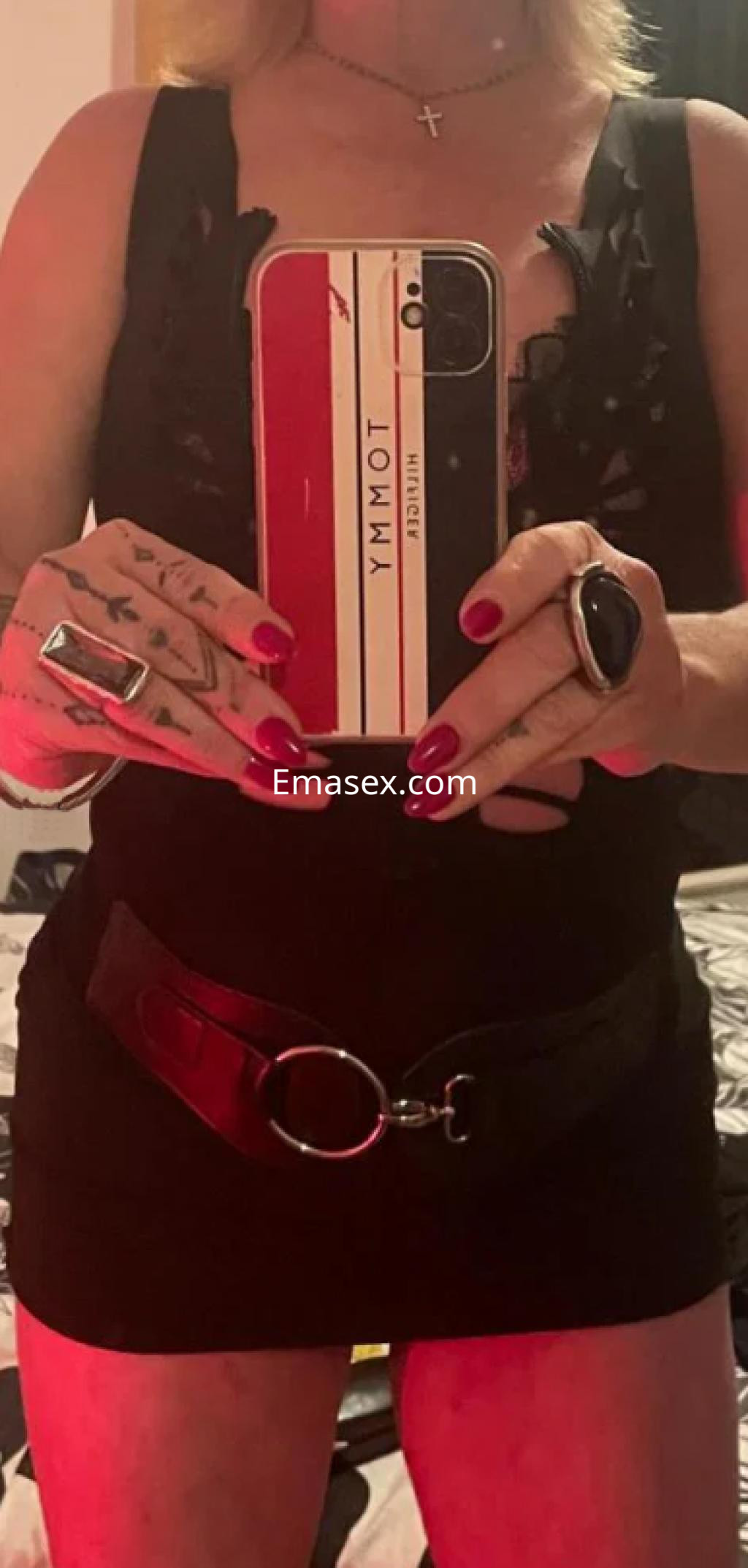 Escort en Girona con cabello rubio y largo, luce un vestido negro ajustado y accesorios llamativos mientras se toma una selfie.