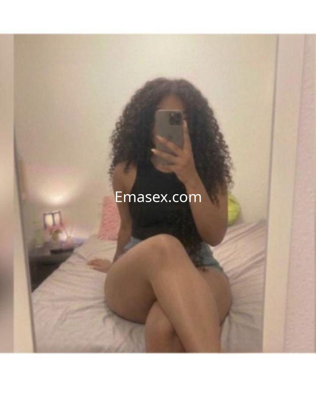 Escort en Barcelona con cabello rizado, sentada en una cama, vistiendo una camiseta negra y pantalones cortos.