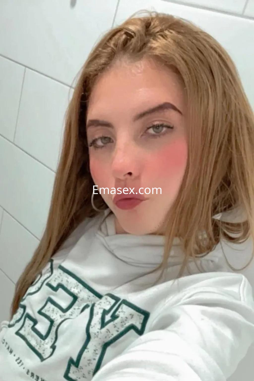 Puta con cabello largo y rubio, viste sudadera blanca y posa con un suave maquillaje en un baño de Cádiz.