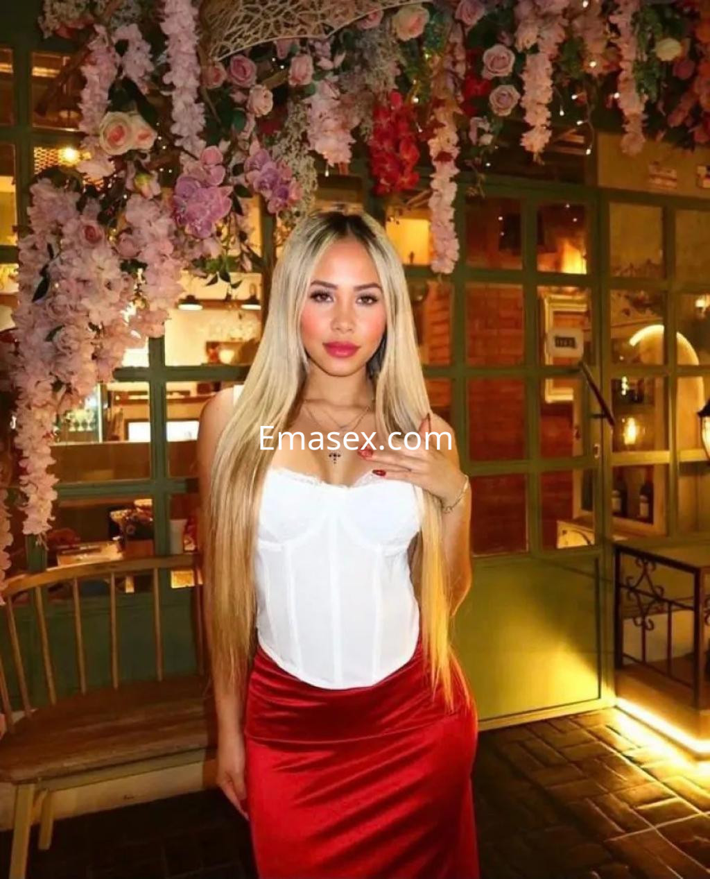 Escort en Palma de Mallorca, con cabello largo y rubio, viste un corset blanco y una falda roja en un entorno floral.