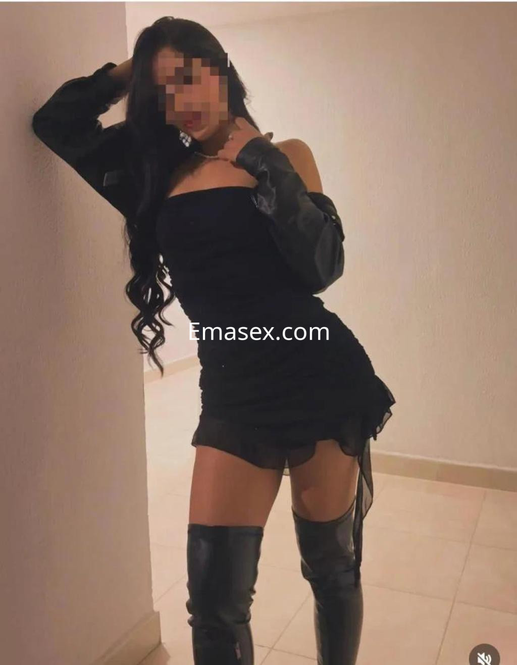 Escort en Valencia posando con un vestido negro ajustado y botas altas, con cabello largo y ondulado.