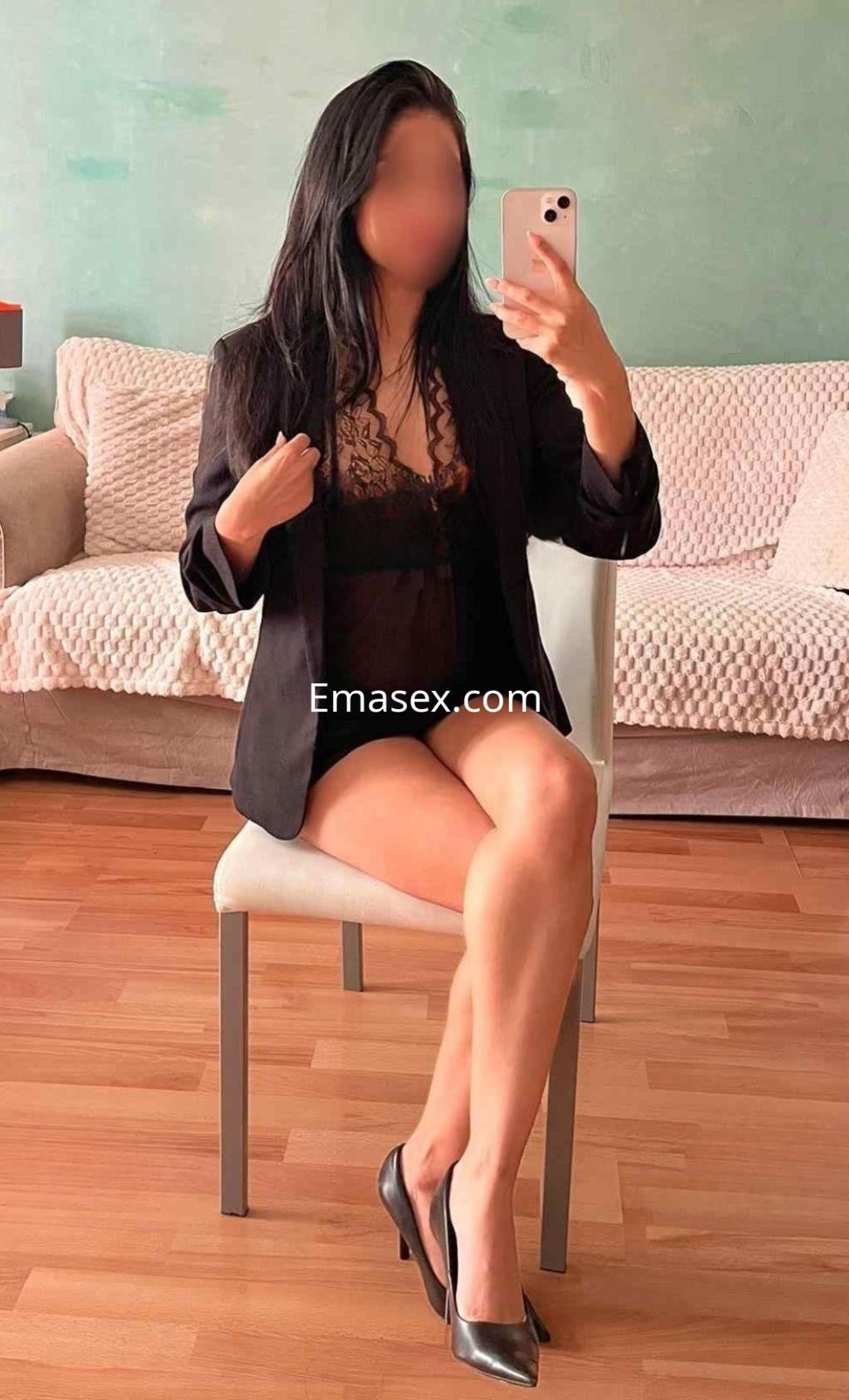 Puta en Madrid sentada en una silla, viste una blusa negra de encaje, blazer y tacones altos en un ambiente acogedor.