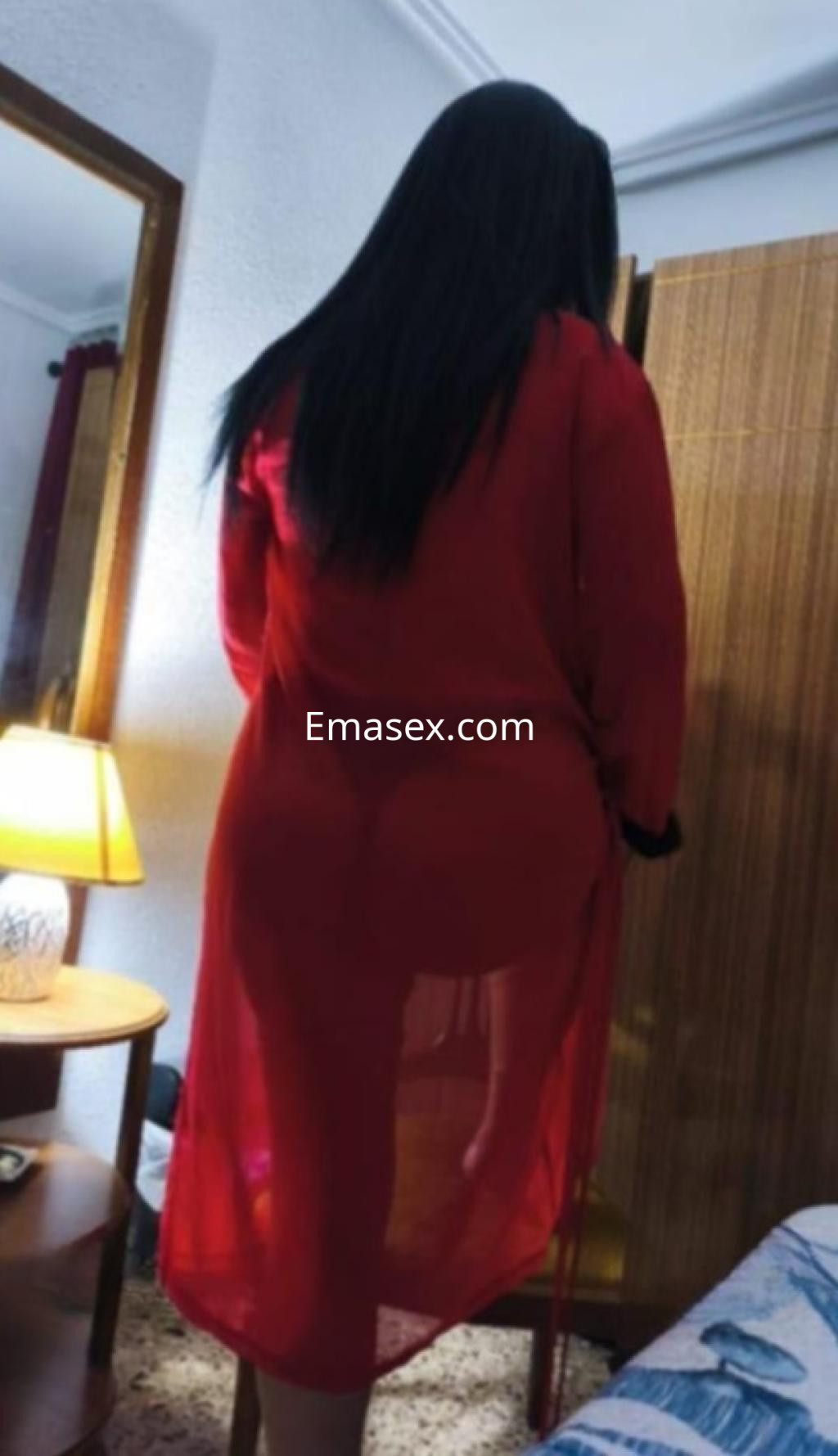 Escort en Valencia, con bata larga roja semitransparente, de espaldas en un ambiente acogedor.