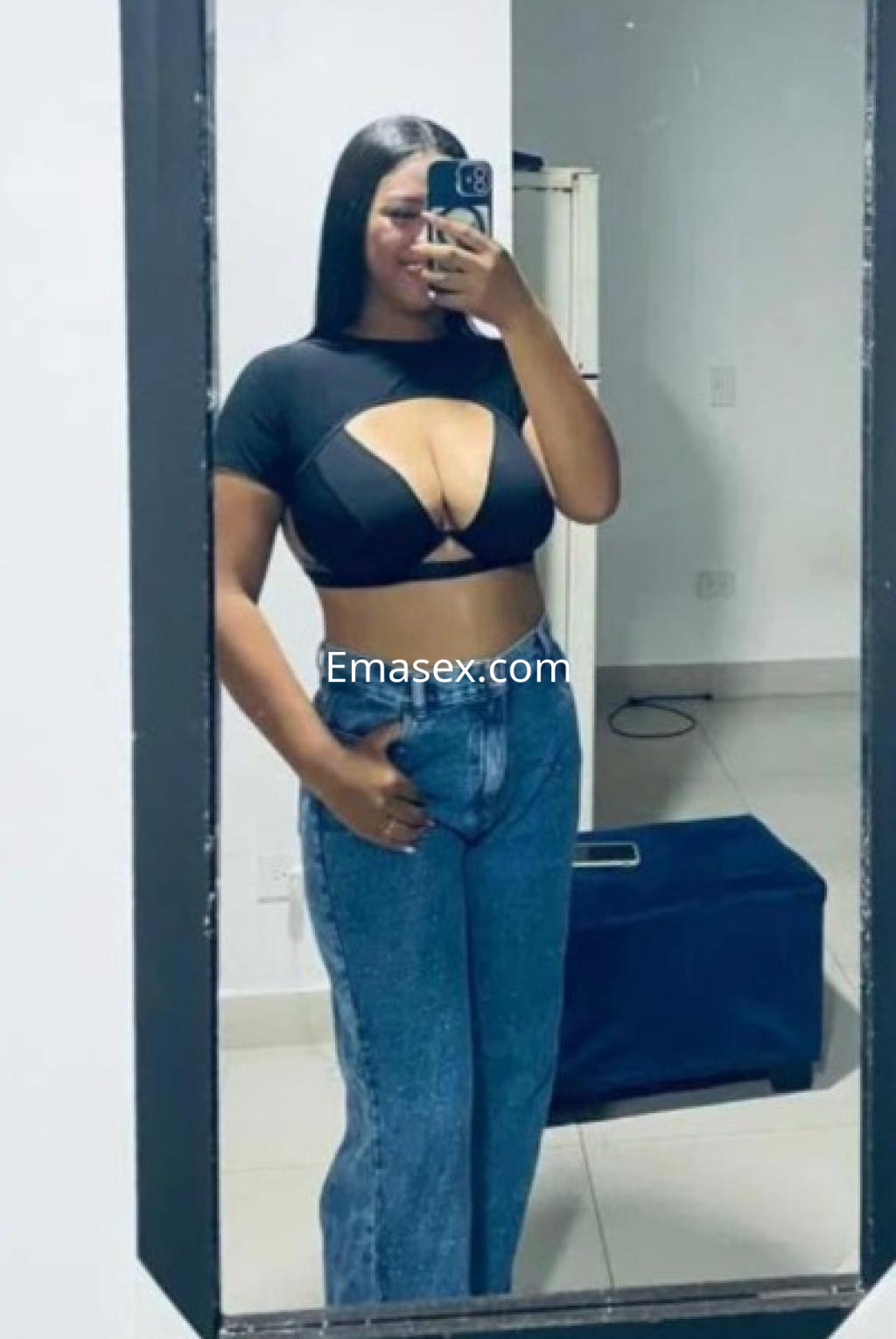 La escort en Girona se toma una selfie en el espejo luciendo un top negro con cortes y jeans ajustados.