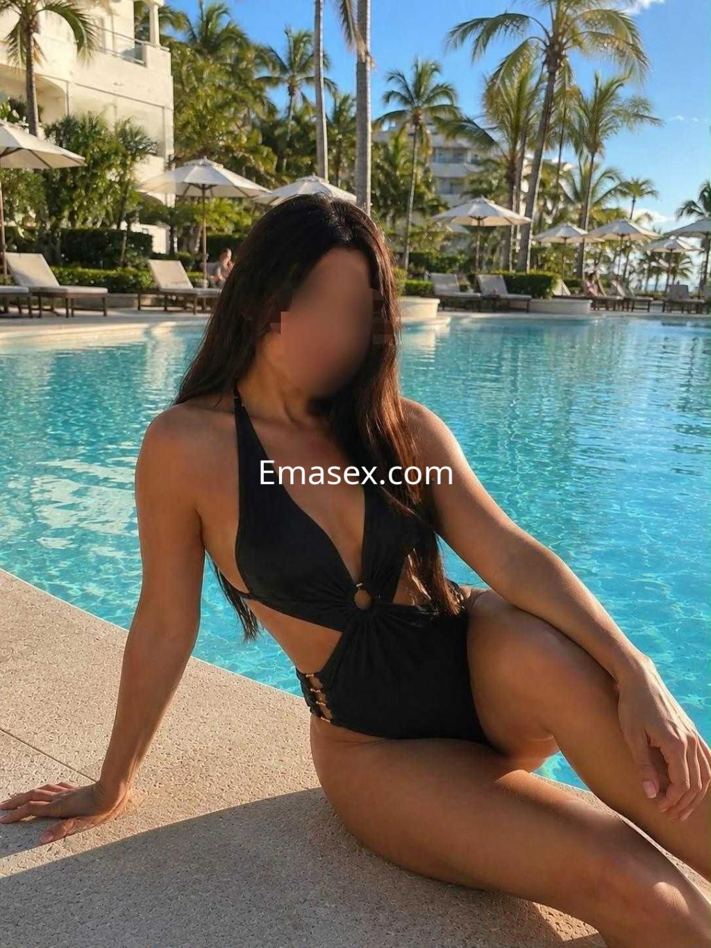 Escort en Cáceres con un traje de baño negro, sentada al borde de una piscina rodeada de palmeras.