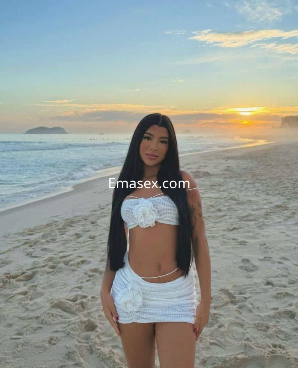 Puta en Málaga, luciendo un atuendo blanco con flores, posa en la playa al atardecer con el mar de fondo.