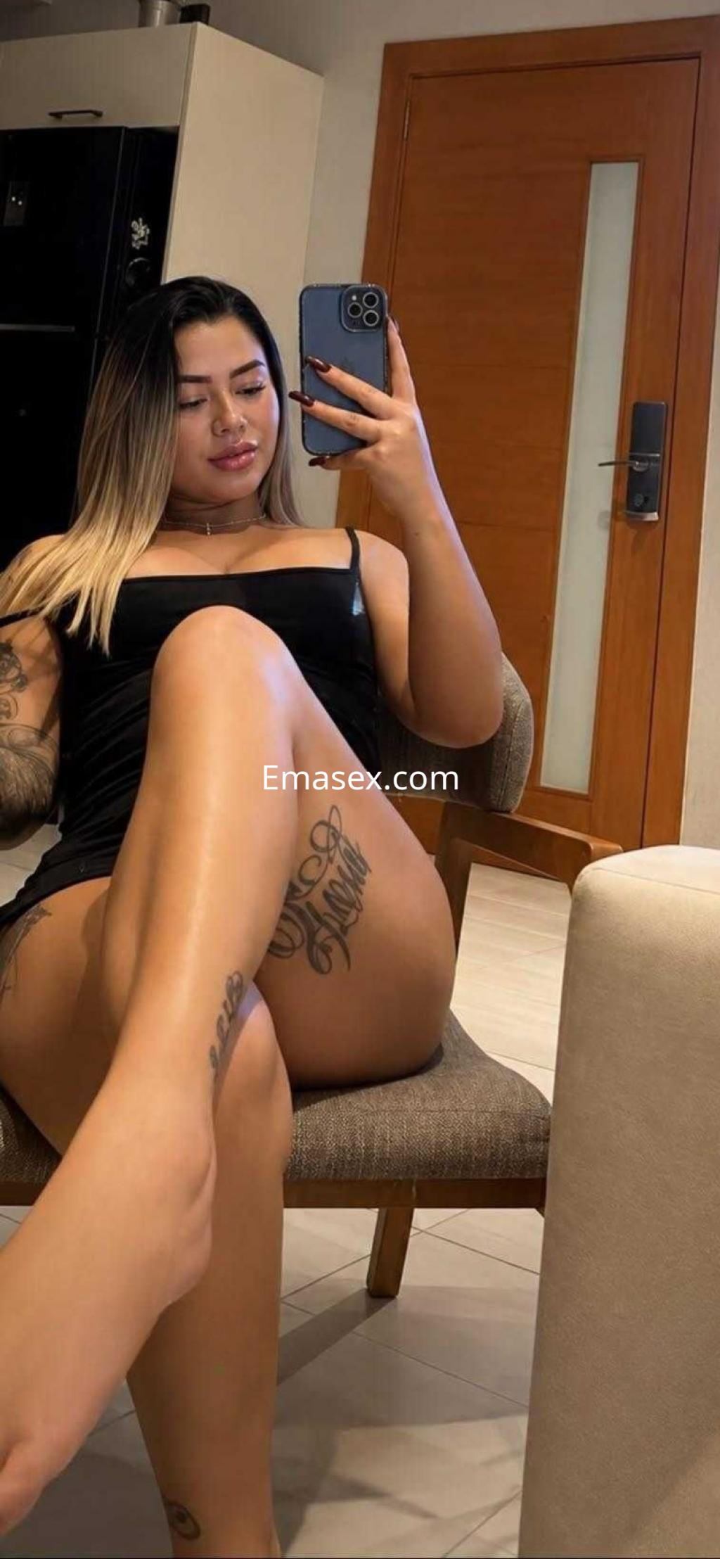Puta en Valencia, sentada en una silla con un vestido negro ajustado, mostrando sus tatuajes y sonriendo a la cámara.