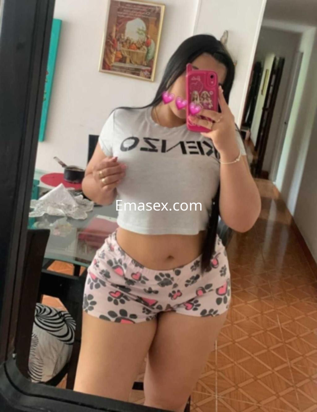 Puta en Alicante viste una camiseta corta gris con texto y pantalones cortos de estampado floral mientras se mira en un espejo.