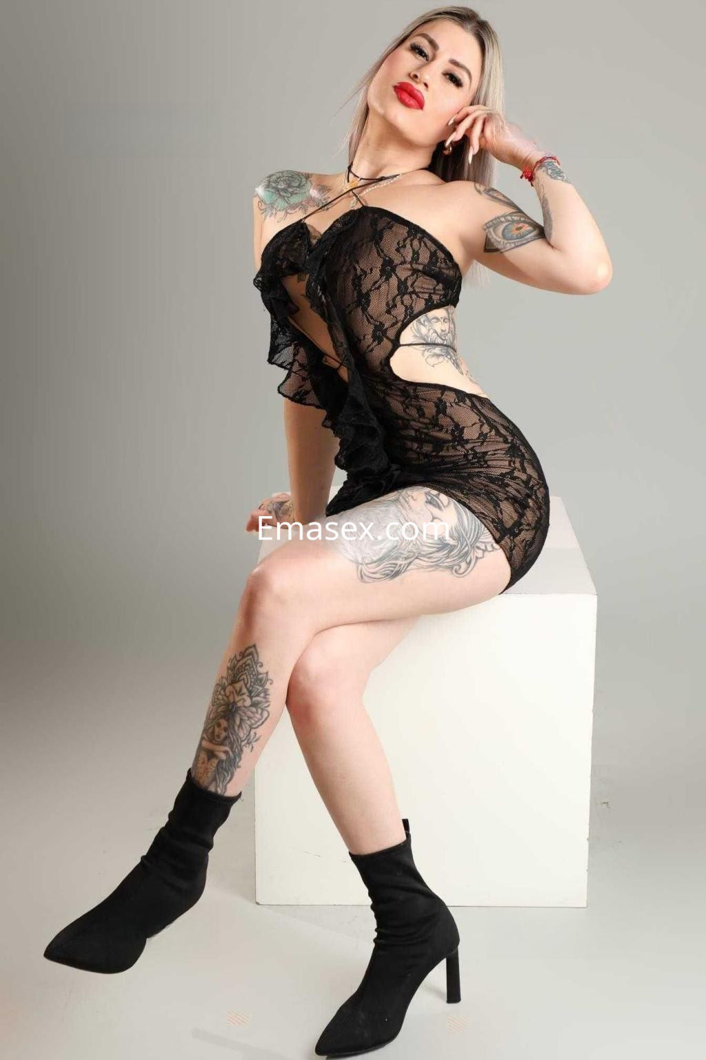 Puta en Zaragoza sentada sobre un pedestal, vistiendo un vestido de encaje negro y mostrando sus tatuajes.