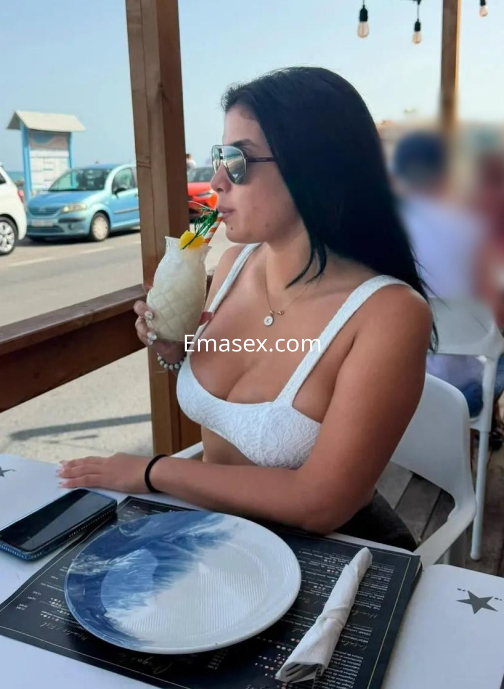 La escort se encuentra en un restaurante al aire libre en Córdoba, con un top blanco, bebiendo de una piña colada.