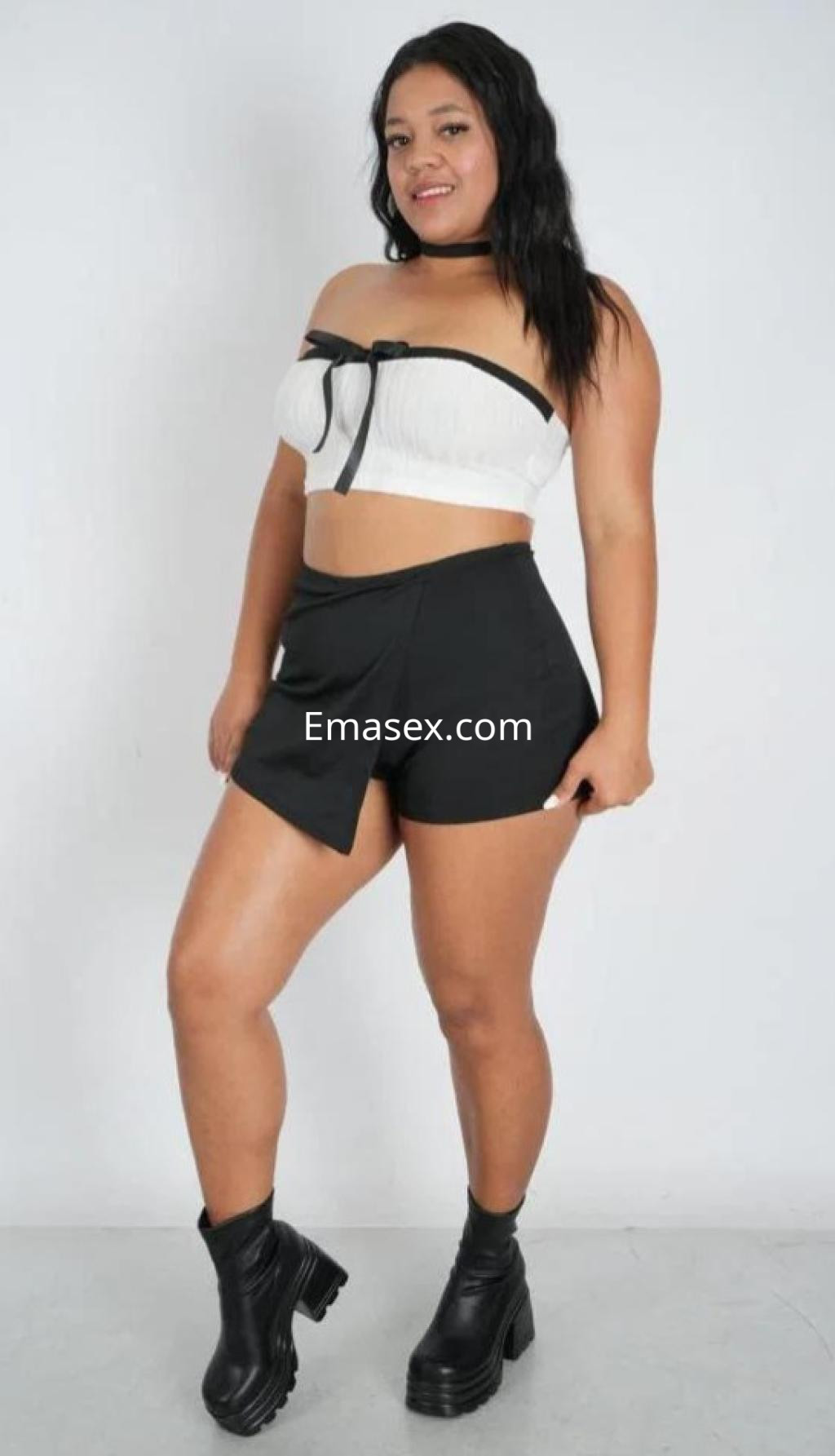 Puta en Alicante, viste un top blanco sin mangas y pantalones cortos negros, posando con una sonrisa y botas.