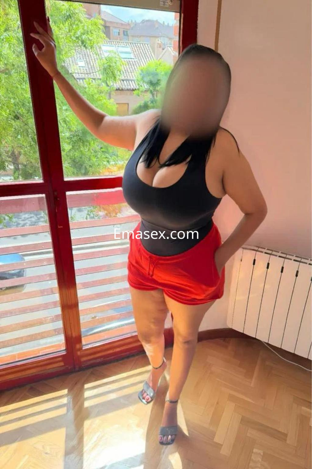 Escort en Toledo posa de pie junto a una ventana, vistiendo un top negro y pantalones cortos rojos.