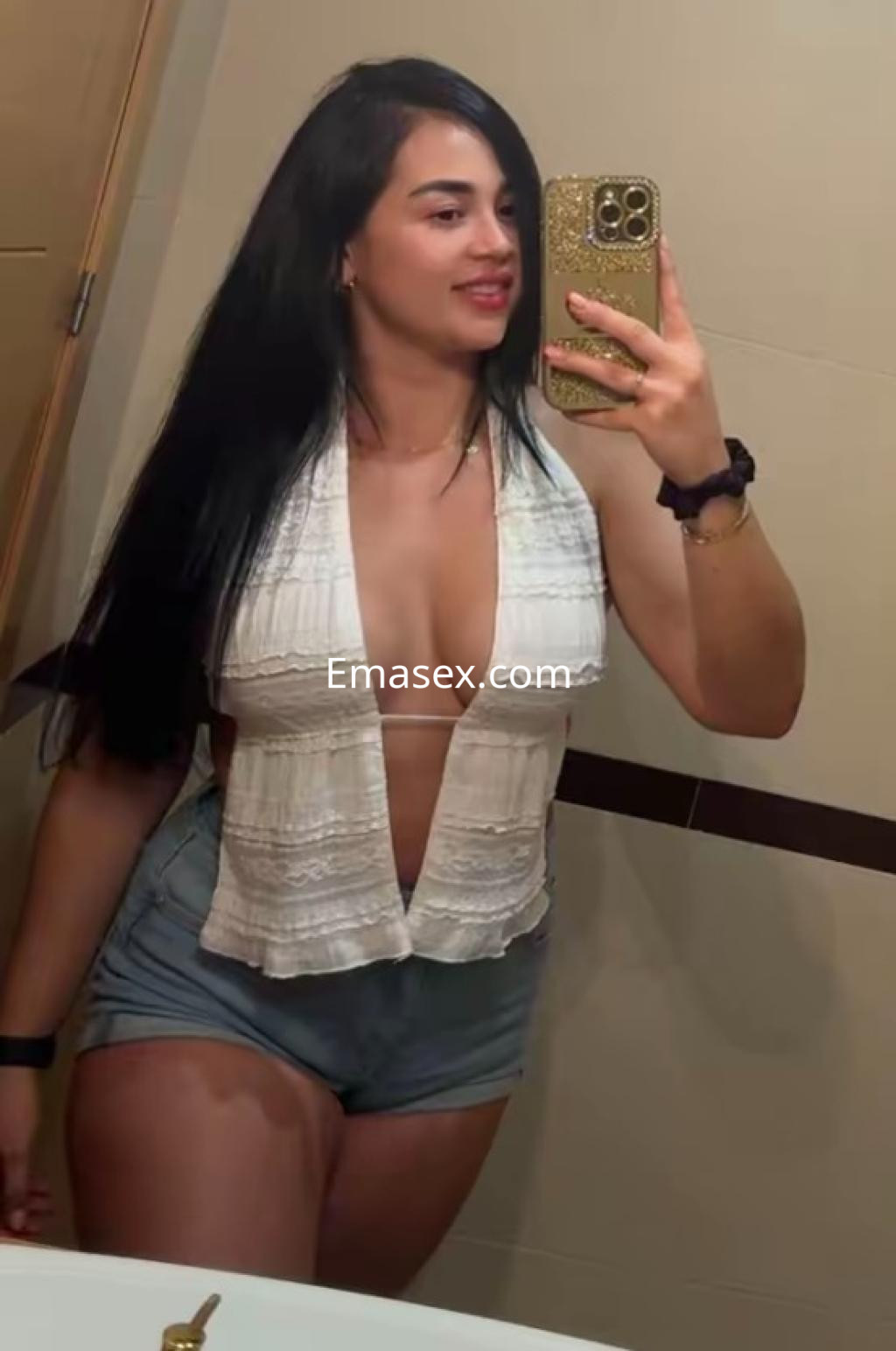 Escort en Murcia posa frente a un espejo, vistiendo una blusa blanca con escote y shorts de mezclilla, sonriendo.