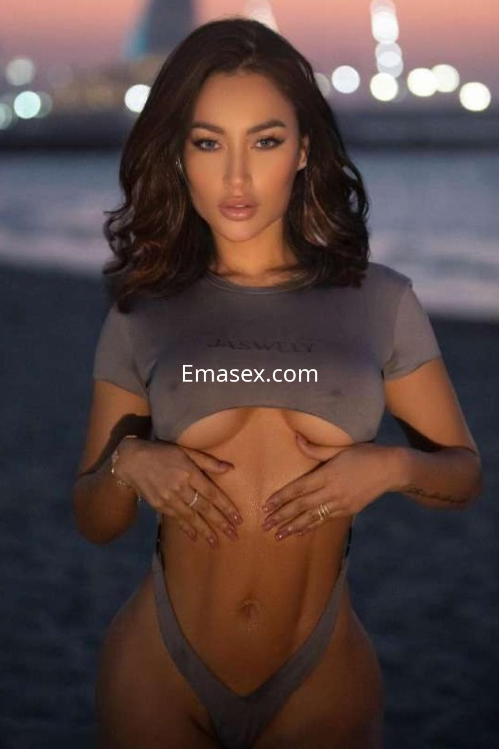 Escort en Ibiza, luciendo un top gris y una lencería mínima, con un fondo de playa al atardecer.