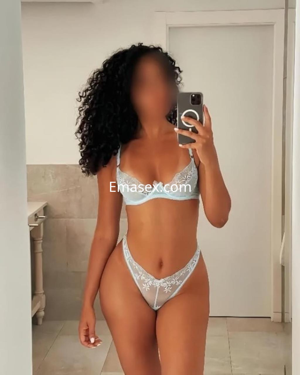 Puta en Girona posando con lencería azul clara en un baño moderno, mostrando su figura y cabello rizado.