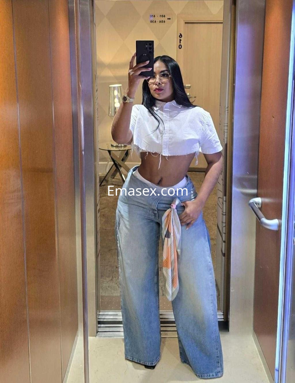 Escort en Badajoz luciendo una blusa corta blanca y pantalones de mezclilla anchos en un ascensor moderno.