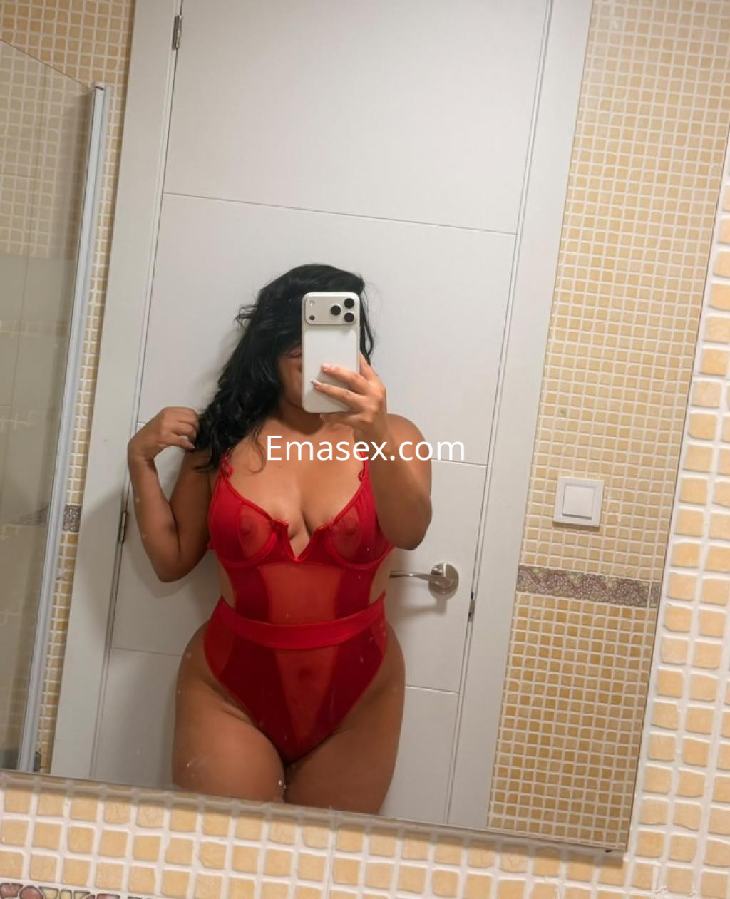 Escort en Castellón posando frente al espejo con un body rojo transparente y cabello oscuro.