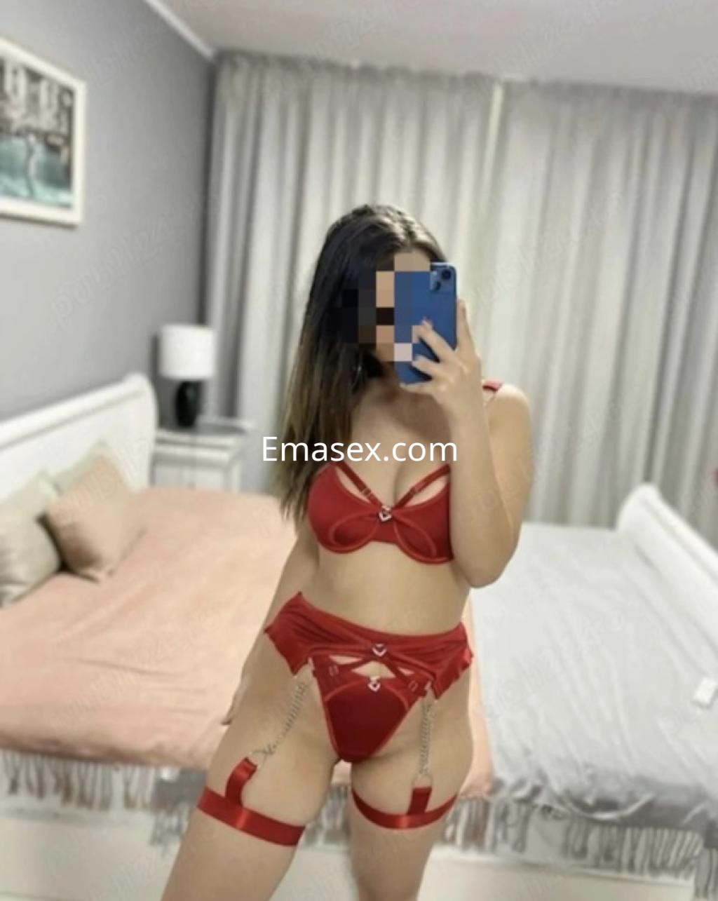 Escort en Castellón posando en un dormitorio, vistiendo lencería roja con detalles brillantes y tirantes.
