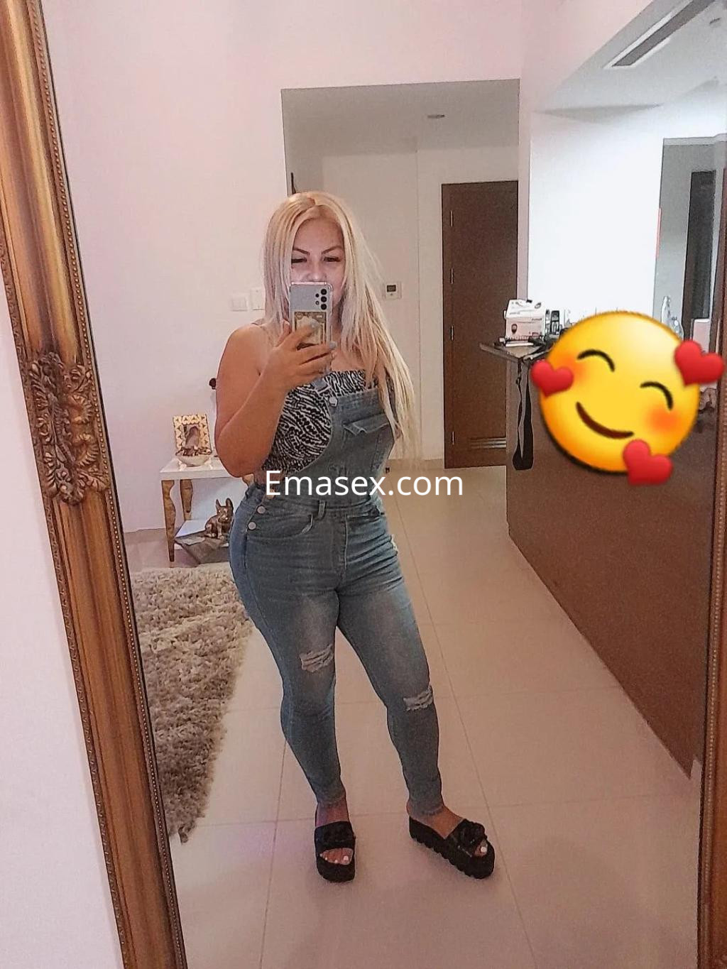 La puta en Murcia lleva un overol de mezclilla ajustado, con blusa de tirantes y sandalias, posando frente al espejo.