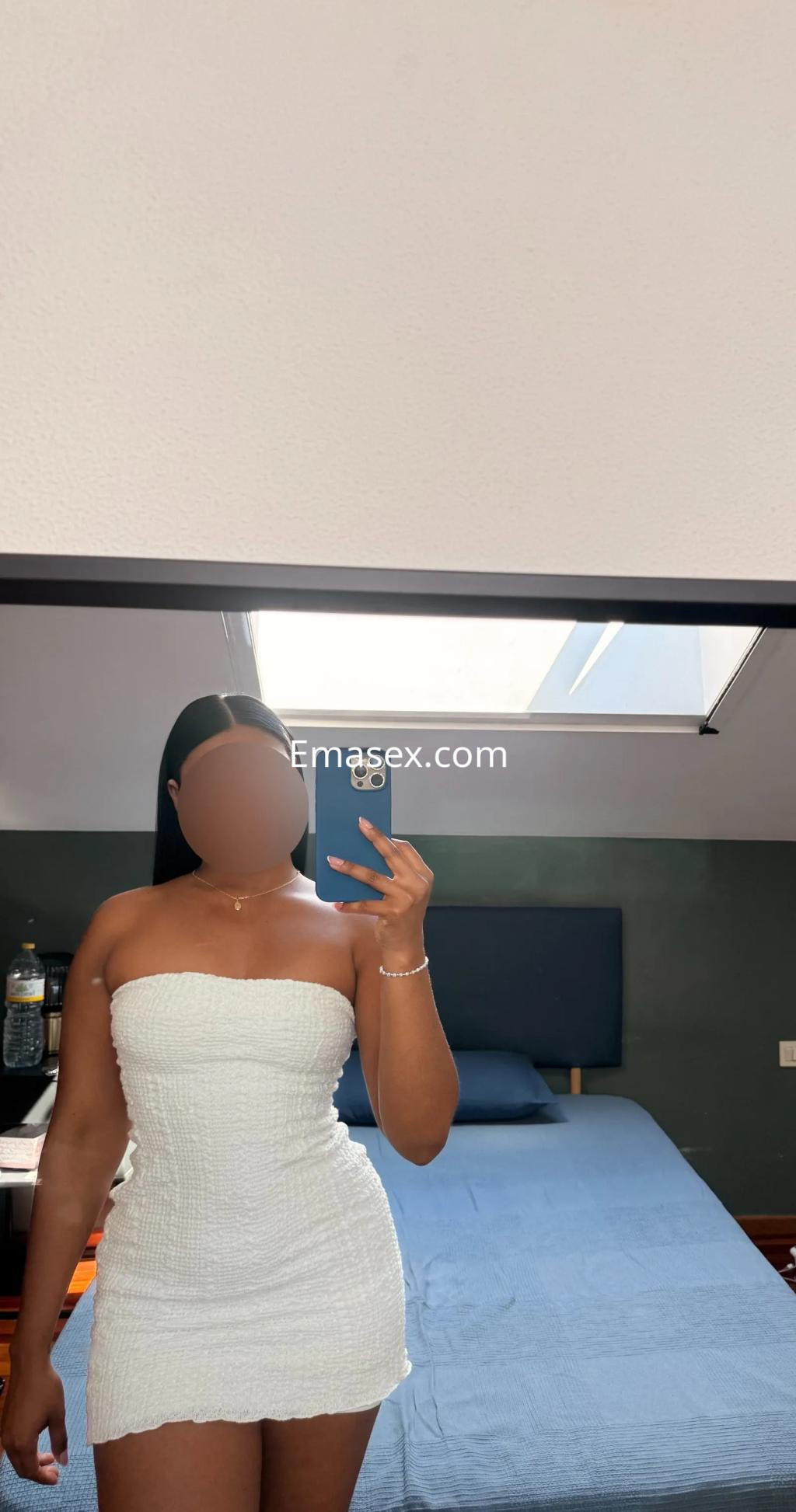 Puta en Vigo luciendo un vestido blanco ajustado mientras se toma una selfie en un dormitorio con cama azul.